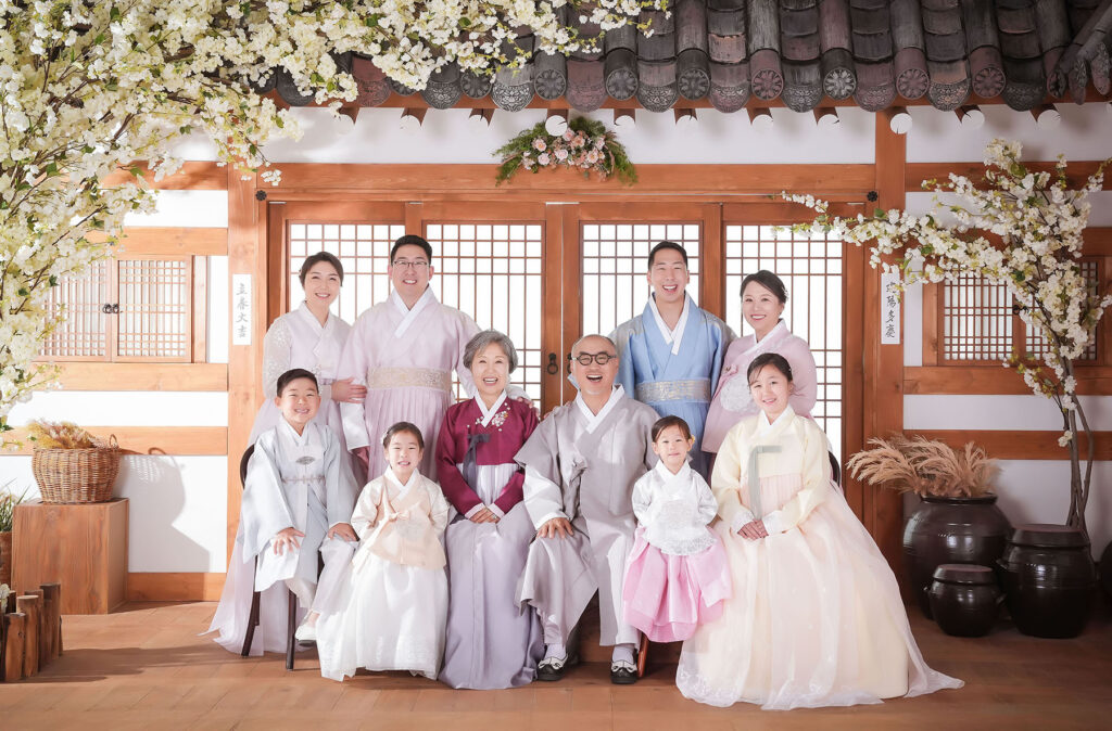 전통 한옥 스튜디오 세트에서 고운 한복을 차려입고 촬영한 대가족 가족사진. 조부모님, 부모님, 자녀들과 손주들까지 여러 세대가 함께 화목한 미소를 짓고 있는 스튜디오 컷